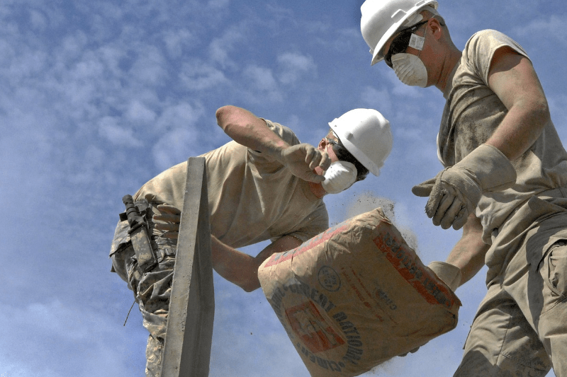 pracownicy budowlani w białych kaskach i maskach przeciwpyłowych rozładowujący worki z cementem, beton