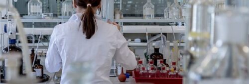 Kobieta ubrana w biały fartuch odwrócona plecami na tle laboratorium wypełnionego szklanymi buteleczkami oraz pipetami