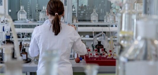 Kobieta ubrana w biały fartuch odwrócona plecami na tle laboratorium wypełnionego szklanymi buteleczkami oraz pipetami