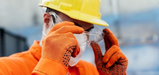 Mężczyzna ubrany w pomarańczowy kombinezon, maseczkę, żółty kask oraz rękawice robocze zakładający okulary ochronny, Ochrona przed substancjami niebezpiecznymi