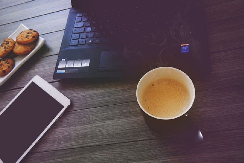 laptop stojący na drewnianym biurku w towarzystwie telefonu, ciasteczek oraz kawy, pozycjonowanie stron