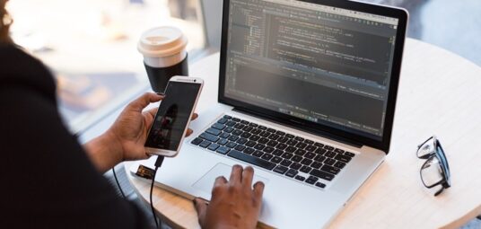 pracujący programista nad front-endem, w ręce trzymający telefon, w tle widać stojący kubek z kawą na wynos oraz okulary