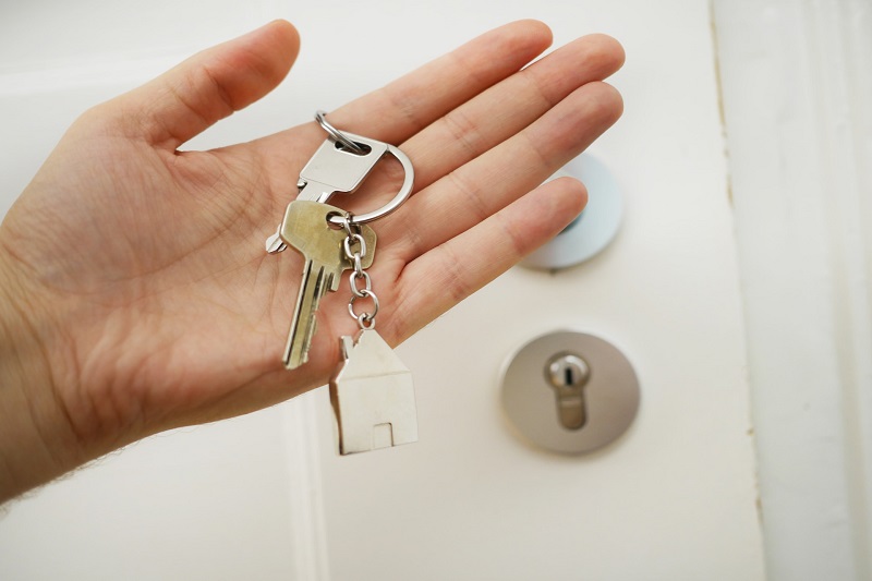 dłoń trzymająca klucze z przypiętym brelokiem w kształcie domu na tle białych drzwi, prawnik