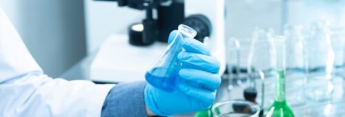 Laboratorium, ręka w nylonowej rękawiczce trzymająca menzurkę napełnioną niebieskim płynem, w tle stojący mikroskop