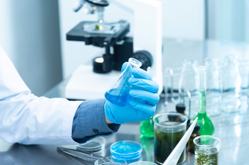 Laboratorium, ręka w nylonowej rękawiczce trzymająca menzurkę napełnioną niebieskim płynem, w tle stojący mikroskop