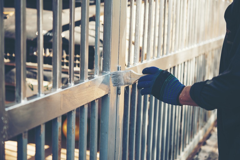 Ręka w ochronnej rękawiczce malująca ogrodzenie z metalowych prętów na srebrny kolor, zabezpieczenie antykorozyjne